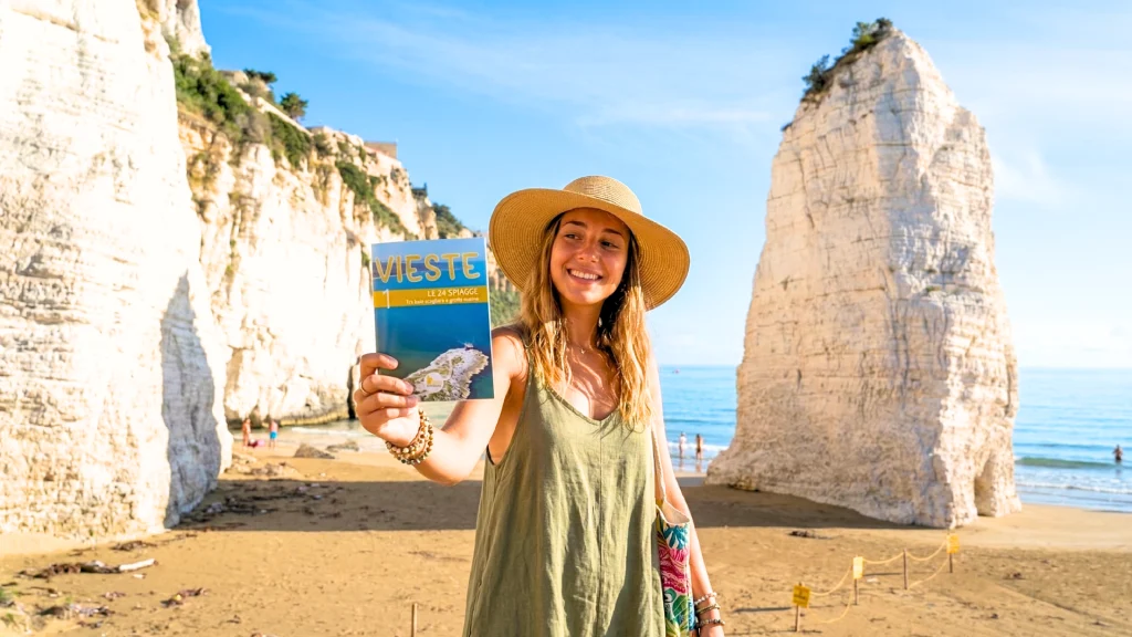 Una ragazza con la guida n.1 di vieste experience in mano, Le 24 spiagge di Vieste. Lei si trova in spiaggia davanti al Pizzomunno di Vieste