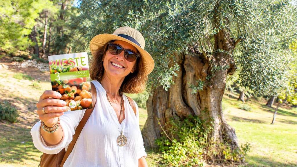 guida n.3 I Sapori di Vieste della collana editoriale di Vieste Experience. Signora ha in mano la guida e alle spalle un albero di Ulivo Secolare